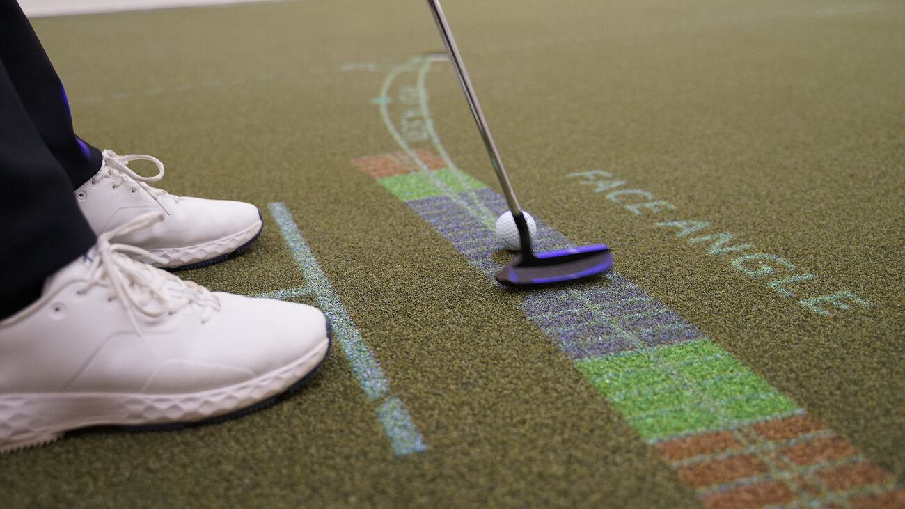 A putter set behind the ball on a colour-banded guide projected on the green, with the words Face Angle and a stance line at the golfer's feet