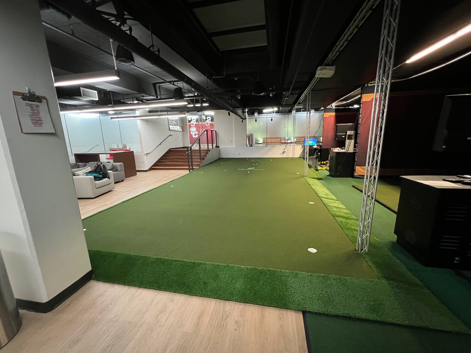 USC golf facility: a wide view of the TOURPUTT putting green, with a lounge and stairs to one side and a simulator bay to the other