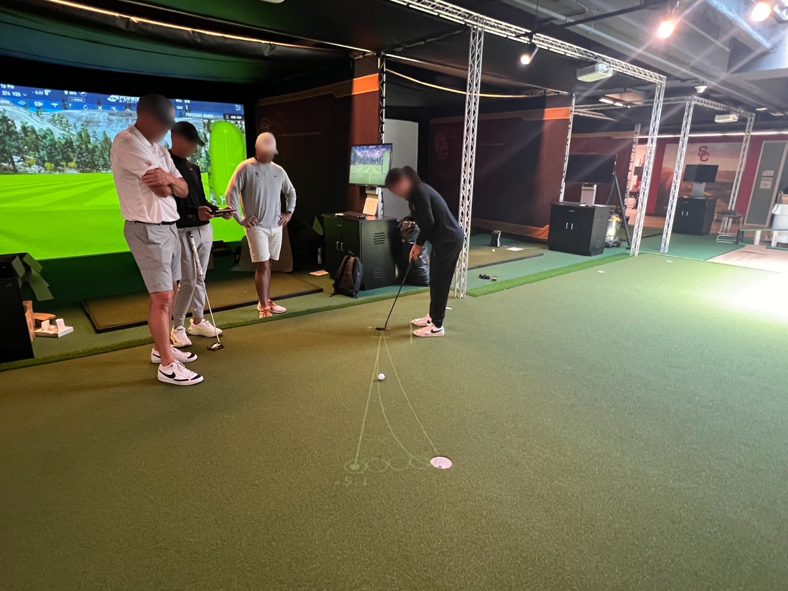 USC golf facility: a golfer putts along projected lines on the TOURPUTT green while three people watch, with the simulator bay behind them. Faces are blurred.